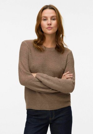 Tan strikket sweater med lange ærmer, rund hals og ribbede manchetter. Parret med mørke jeans, der fremhæver et tekstureret stof.