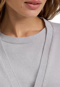 Maglione grigio con scollo a V, texture lavorata a maglia e dettagli di cucitura sottili lungo i bordi. Focus sul colletto e sulla qualità del tessuto.