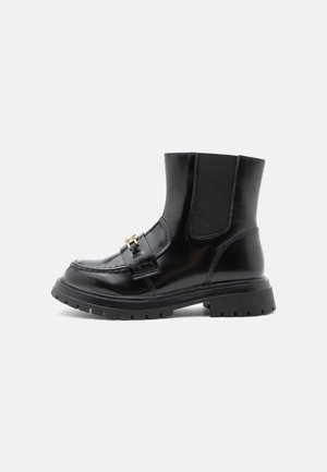 Botas de tornozelo em pele preta com sola robusta, painéis laterais elásticos, costura moc-toe e fivela decorativa dourada em fundo branco.