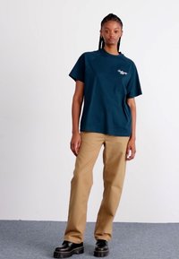 Teal cotton t-shirt with short sleeves and a logo on the left chest, paired with beige pants and black platform shoes. Plain background.