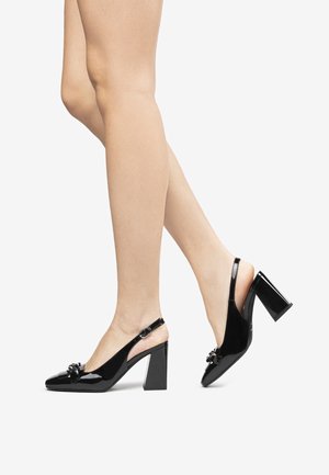 Tacones slingback de charol negro con tacones gruesos, usados en piernas cruzadas contra un fondo blanco liso.
