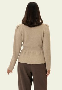 Beiger, gerippter Pullover mit langen Ärmeln, tailliertem Schnitt und Rüschen-Details. Weiche Textur und dezente elastische Akzente betonen die Form.