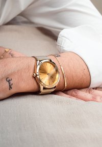 Montre-bracelet en or avec un cadran rond, un cadran en or clair, des index horaires fins et un bracelet en métal, portée sur un poignet avec un bracelet en or.