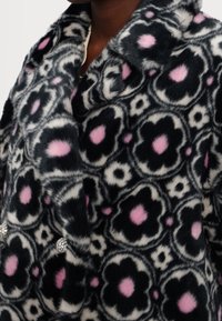 Cappotto in pelliccia sintetica con motivo floreale in nero, bianco e rosa. Decorato con bottoni di strass. Texture morbida con colletto a rever.
