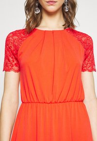 Czerwona sukienka z koronkowymi krótkimi rękawkami, plisowanym przodem i elastyczną talią. Miękka tkanina o płynącej fakturze, odpowiednia na codzienne lub półformalnie okazje.