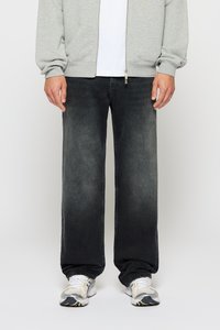 Mann trägt ausgewaschene schwarze Jeans mit weitem Bein, weiße Turnschuhe, weißes Hemd und hellgrauen Reißverschluss-Sweatshirt, steht vor einem schlichten Hintergrund.