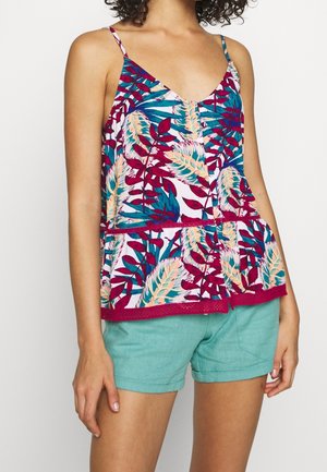 Frau trägt ein ärmelloses Peplum-Top mit bunten Blattmustern und türkisfarbene Shorts, stehend vor einem schlichten Hintergrund.