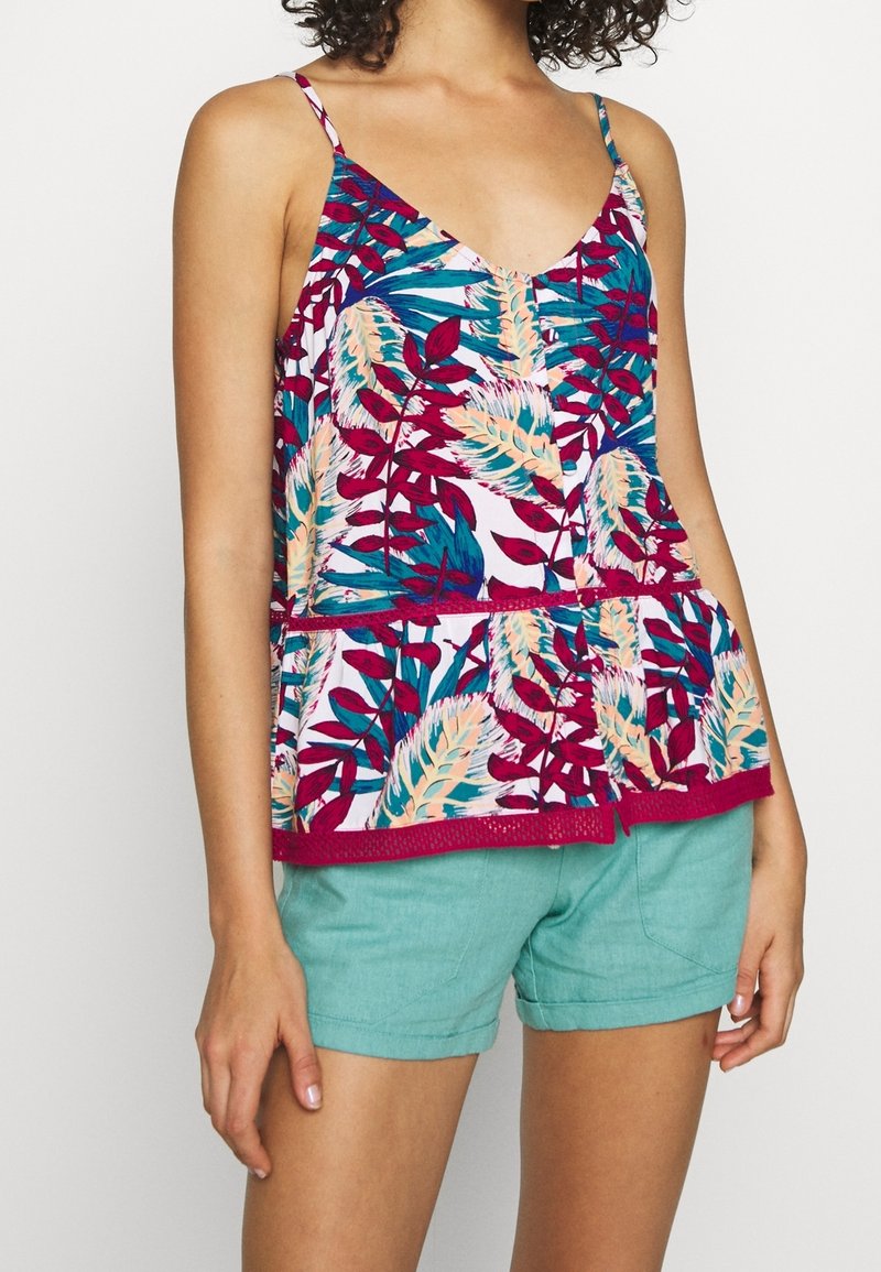 Femme portant un top peplum sans manches avec des motifs de feuilles colorées et un short bleu sarcelle, debout devant un fond uni.