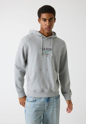 Joven con sudadera con capucha gris claro con el logo "GUESS jeans" y vaqueros azul claro, posando contra un fondo claro y liso.