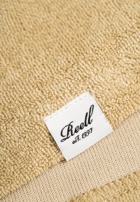 Beige Handtuch mit einer strukturierten, gewebten Oberfläche und einer glatten Unterseite. Enthält ein weißes Etikett mit schwarzem Text: "Reell est. 1997."