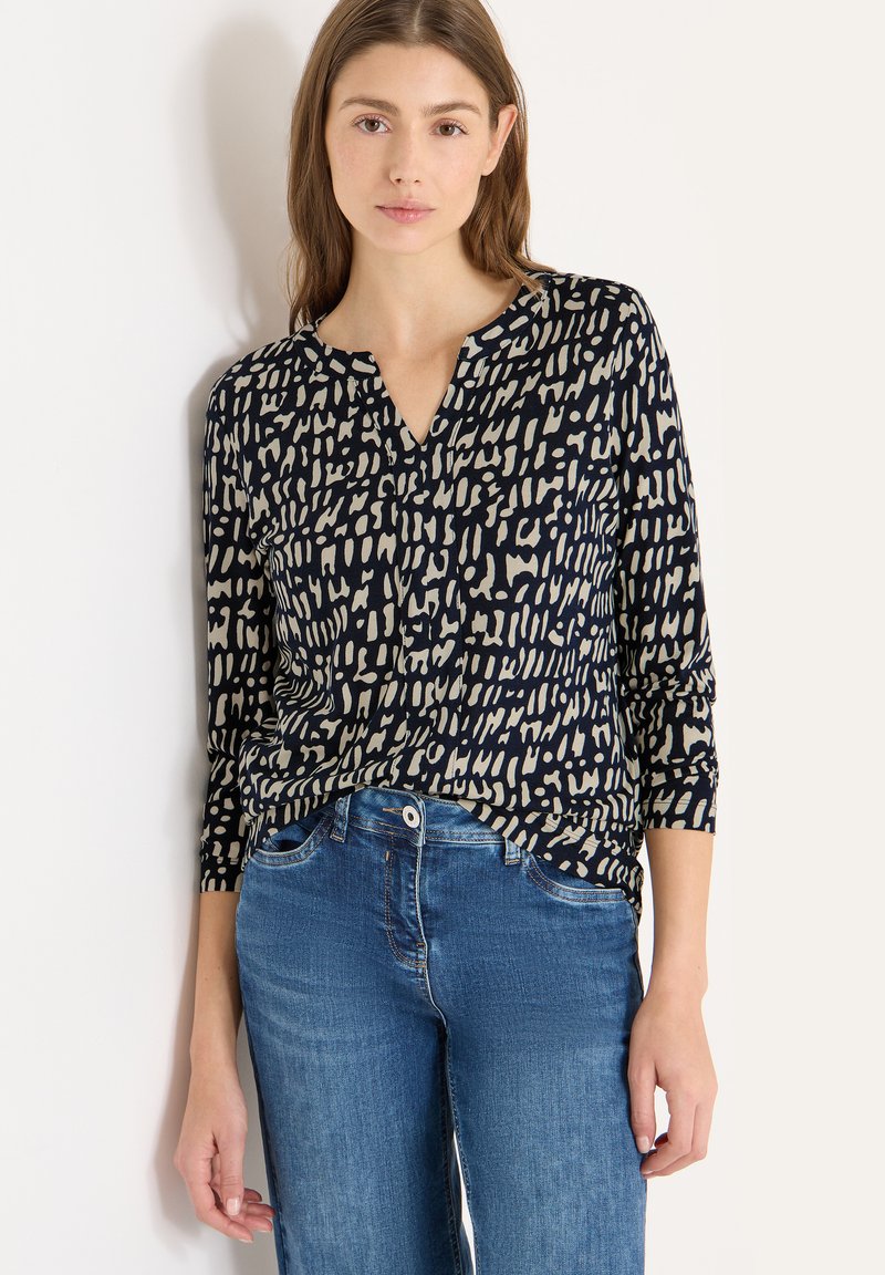 Marineblå bluse med lange ærmer og beige abstrakt mønster, v-udskæring og blødt stof, kombineret med lyseblå jeans. Glat tekstur, afslappet pasform.