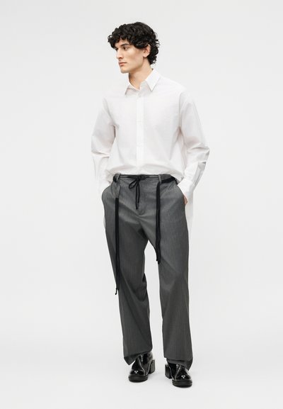 Mann mit lockigem Haar, der ein weißes Hemd mit Knöpfen, graue Hosen mit Nadelstreifen und langen schwarzen Kordeln sowie schwarze Schuhe mit Absatz trägt, steht seitlich.