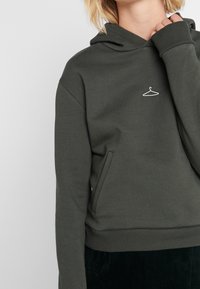 Kápová mikina v olivově zelené barvě s textured povrchem, na hrudi s malým bílým grafickým motivem ramínka a klokaní kapsou.