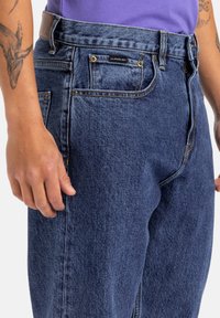 Jeans de mezclilla azul oscuro con un corte clásico, que presentan herrajes dorados y un parche de cuero en la cintura. Tejido texturizado con bolsillos laterales.