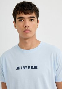 Camiseta de algodón azul claro con mangas cortas y cuello redondo. Presenta un texto azul oscuro que dice "TODO LO QUE VEO ES AZUL" en el pecho.