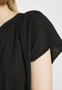 Haut en lin noir à coupe décontractée, présentant des détails froncés au niveau du col et des manches courtes. Texture douce avec une finition légèrement transparente.