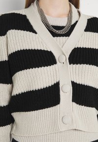 Cárdigan de punto con rayas horizontales en negro y crema, que presenta un escote en V, tres botones redondos y un tejido texturizado. Se ve una gargantilla de cadena plateada.