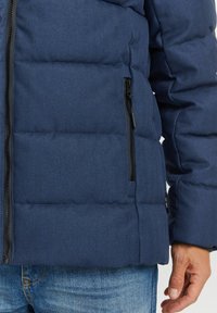 Marineblaue Steppjacke mit Fischgrätmuster, ausgestattet mit einem Frontreißverschluss und einer Seitentasche. Das Material wirkt robust und isoliert.