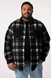 Chemise en flanelle à carreaux noir et blanc avec deux poches poitrine et fermeture à boutons, superposée sur une chemise noire, portée avec un jean bleu clair.