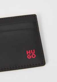 Cardholder din piele neagră cu multiple sloturi. Prezintă logo-ul roșu "HUGO". Cusăturile vizibile de-a lungul marginilor adaugă detalii designului.