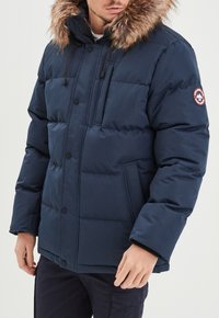 Marineblaue Steppjacke mit fellgefütterter Kapuze, Knopfverschluss, mehreren Taschen und gesteppten Einsätzen. Mit Logo-Patch am Ärmel.