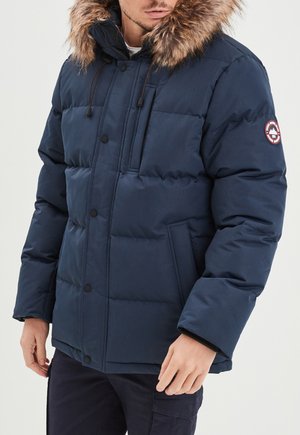Chaqueta acolchada de color azul marino con capucha forrada de piel, cierre de botones, múltiples bolsillos y paneles cosidos. Presenta un parche con el logo en la manga.