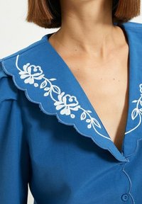 Camicia blu con colletto ondulato e ricamo floreale bianco, dettagli con bottoni sulla parte anteriore e tessuto morbido.