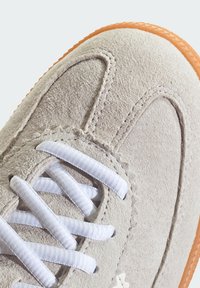 Beige semišové tenisky s bílými tkaničkami, se šitým vzorem a texturovaným povrchem. Gumová podrážka v světlé oranžové barvě.
