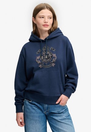 Ung kvinde med skulderlangt hår iført en mørkeblå hættetrøje med en broderet våbenskjold og lyseblå jeans, kigger let mod venstre.