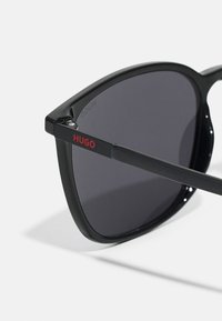 Ochelari de soare negri cu lentile închise, design unghiular, având un logo roșu "HUGO" pe braț și un finisaj elegant, lucios.