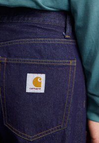 Jean en denim de couleur bleu foncé, avec des coutures jaunes et une étiquette blanche portant un logo jaune. Présenté en gros plan sur la poche arrière.