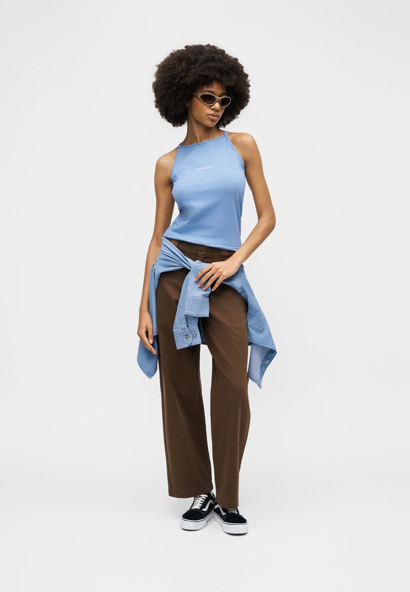 Donna con capelli afro che indossa occhiali da sole, una canotta azzurra, pantaloni marroni, una camicia blu annodata in vita e scarpe da ginnastica nere.