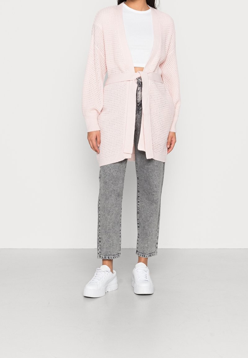 Světle růžový pletený kardigan se zavazováním v pase, otevřený předek a dlouhé rukávy, v kombinaci se šedými džínami s vysokým pasem a bílými teniskami.