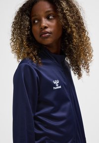 Navy zip-up jacket made of smooth fabric, featuring a white logo on the chest. Curly hair visible, with a focus on the jacket's detail.