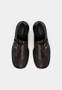 Chaussures en cuir marron foncé à enfiler, avec bouts carrés et boucles en argent, vues de dessus sur un fond clair uni.
