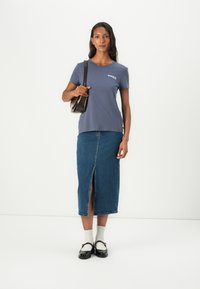 Marineblå T-skjorte med kortermet og logo, kombinert med et kne-langt denimskjørt med en frontsplitt. Modellen bærer en brun skulderveske.