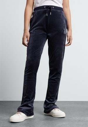 Persoon draagt marineblauwe velours joggingbroek met trekkoord in de taille en witte sneakers, staand op een grijze vloer tegen een effen lichte muur.