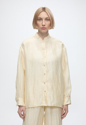 Femme aux cheveux blonds courts portant une chemise ample beige clair à boutons et un pantalon assorti, debout devant un fond blanc uni.