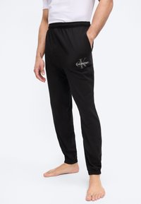 Άνδρας που φοράει μαύρα παντελόνια jogger Calvin Klein με ελαστική μέση και μανσέτες, το ένα χέρι στην τσέπη, ξυπόλυτος σε απλό λευκό φόντο.