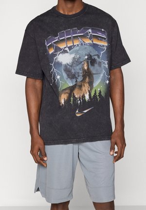 T-shirt noir Nike avec un loup hurlant à la pleine lune, des éclairs, des arbres et le logo Nike, porté avec un short gris clair.
