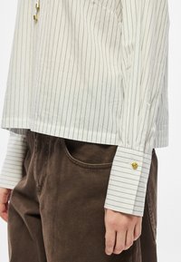 Biała koszula w paski zapinana na guziki, z długimi rękawami, złotymi guzikami i luźnym krojem. W zestawie z brązowymi spodniami sztruksowymi.