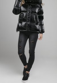 Czarna, błyszcząca kurtka puchowa z kapturem, z pikowanymi sekcjami i przednimi kieszeniami. W zestawie z zniszczonymi czarnymi dżinsami i czarnymi sneakersami.