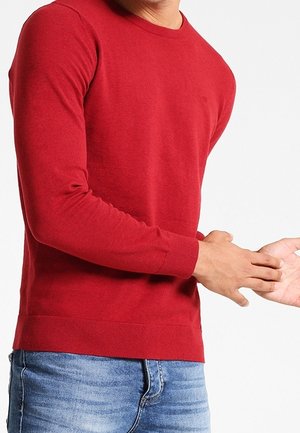 Man wearing a red long-sleeve sweater and blue jeans adjusting his right sleeve against a plain white background.