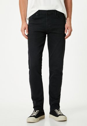 Schwarze Denim-Jeans im Slim-Fit, mit Fünf-Taschen-Design, Gürtelschlaufen und geradem Bein; kombiniert mit schwarz-weißen Sneakers.