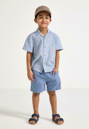 Ung dreng står og smiler iført en lyseblå skjorte med korte ærmer og knapper, matchende shorts, beige kasket og marineblå sandaler.