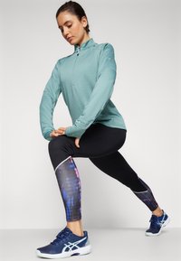 Haut de sport à manches longues en bleu sarcelle clair avec fermeture éclair quart, associé à un legging noir présentant un motif coloré bleu et violet, et des chaussures de sport navy.