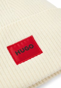 Berretto a maglia color crema con una finitura testurizzata. Presenta una prominente etichetta rettangolare rossa con la scritta "HUGO" in nero.