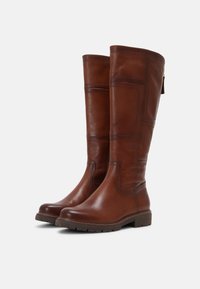 Une paire de bottes en cuir marron hautes avec des talons bas, des bouts ronds et des détails cousus, présentée sur un fond blanc uni.