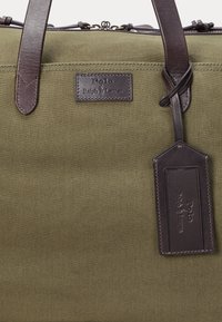 Olijfgroene canvas duffel tas met bruine leren handvatten en accenten. Voorzien van een leren label met reliëflogo en gestikte details.