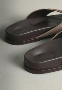 Tongs marron avec une semelle en caoutchouc épaisse et des brides larges et arrondies. La surface est lisse avec un marquage discret sur la semelle intérieure.